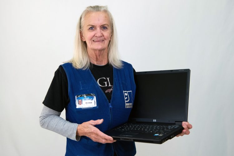 Roslind, a Goodwill team member in a blue uniform, smiles while holding a refurbished laptop from STUG in front of a white studio background.